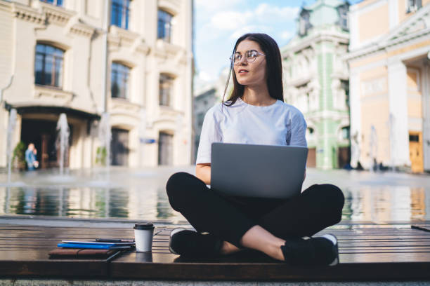 istock Contemplative female student in classic eyeglasses thoughtful looking away while e learning on distance using modern laptop computer during digital travelling, pondering hipster girl with netbook 1421919928