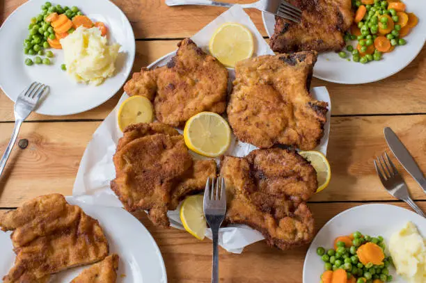 Homemade dinner or lunch table with fried and breaded pork chops, mashed potatoes and buttered peas and carrots. Served on wooden background. Overhead view