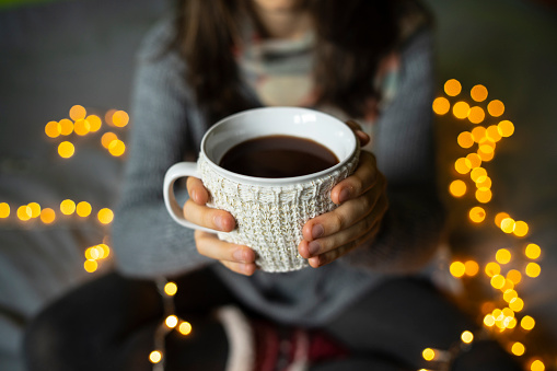 A white mug in a white  cup-cozy held by a pair of hands of an unrecognizable person with a backdrop of unfocussed fairy lights