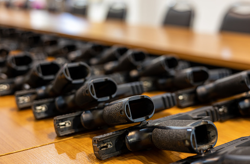 A close-up view of a type of black automatic carbine lined up in a long line on a wooden table in the conference room awaiting a supervisor's inspection.