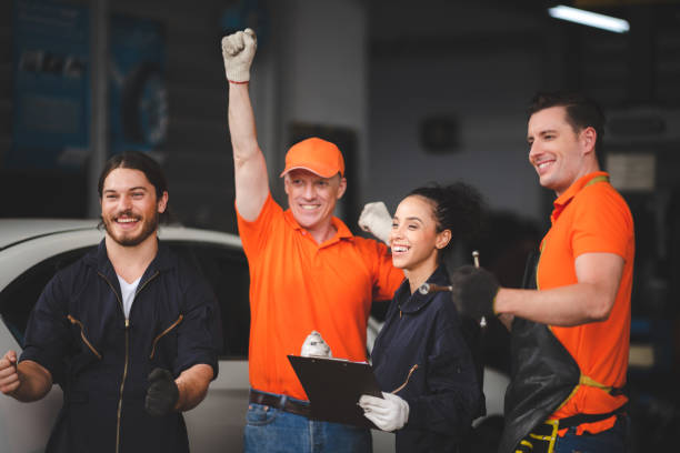 Group of young and senior male and female car mechanics in garage wearing uniform enjoying and celebrating with female worker holding clipping board stock photo