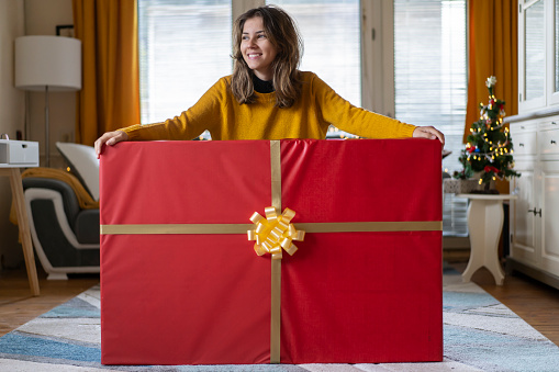 Portrait of happy brunette young woman in santa hat and checkered shirt standing throwing gift boxes in air or catching presents, christmas shopping. indoor studio shot isolated on orange background