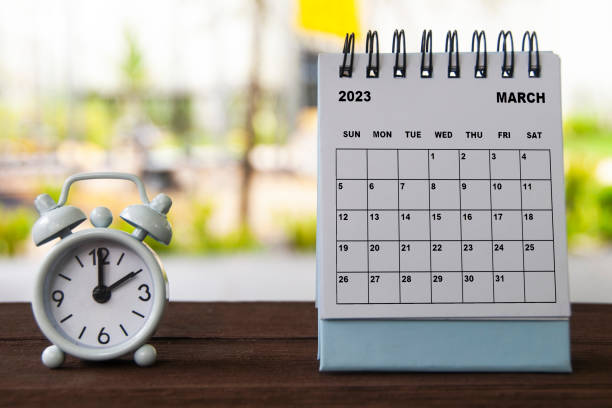 istock Alarm clock pointing at 2 o'clock with March calendar on a bench. Time and calendar concept 1421942522