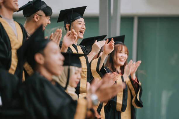 Asian diversify students in graduation gown clapping hands cheering for classmate during graduation ceremony outdoor stock photo