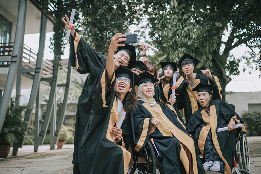 Proud Asian college students in graduation gown selfie with phone outside college buildings