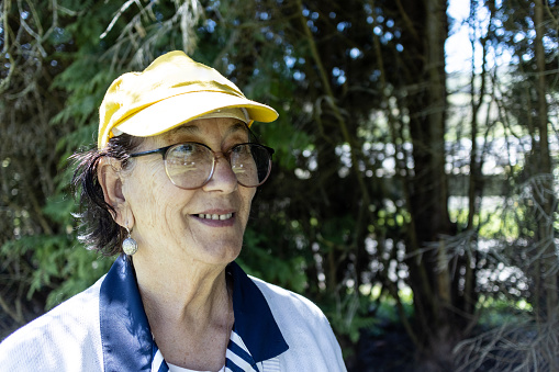 Portrait of a senior woman smiling standing outdoors wearing a cap and eyeglasses looking away with trees in the background.