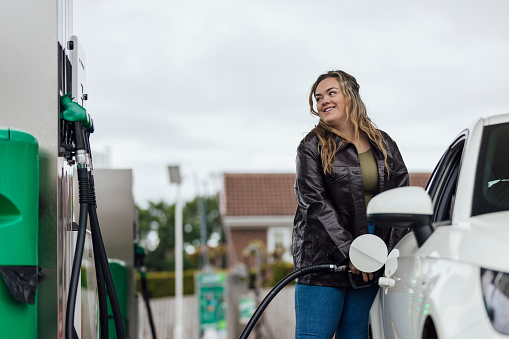 Woman filling her car at a petrol station in the North East of England. Image taken during a cost of living crisis. She is smiling, looking at the car pump screen.