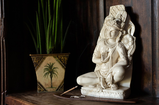 A steaming aroma stick is on a stand against a background of porcelain statue of the Indian god and a green flower in a pot.