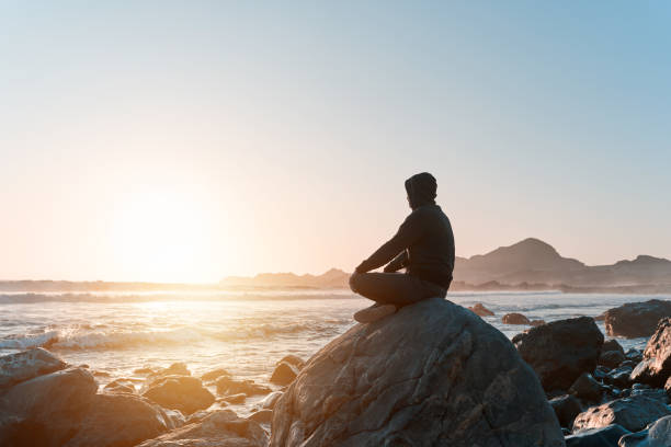 silhouette of a person sitting meditating on the rock on the coast at sunset stock photo