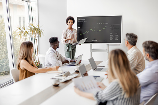 Black Businesswoman Gives Report/ Presentation to Her Business Colleagues in the Conference Room, She Shows Graphics and Company's Growth on the Wall TV