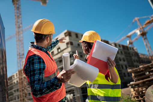 Male and female workers looking at checklist of daily jobs, talking about work schedule with construction site of a multi-story apartment building in background