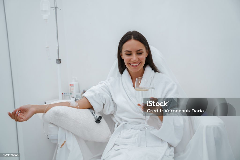 Beautiful woman in white bathrobe drink water during medical procedure in beauty clinic Woman in white bathrobe drink water during medical procedure in beauty clinic. High quality photo IV Drip Stock Photo