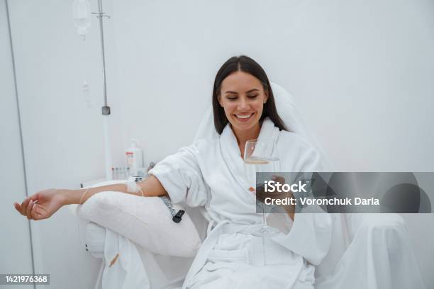 Beautiful Woman In White Bathrobe Drink Water During Medical Procedure In Beauty Clinic Stock Photo - Download Image Now