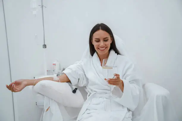 Photo of Beautiful woman in white bathrobe drink water during medical procedure in beauty clinic