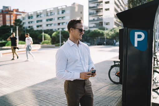Caucasian businessman in trendy sunglasses ticketing in parking meter during time in financial district of megalopolis, formally dressed male manager checking security payment at terminal machine