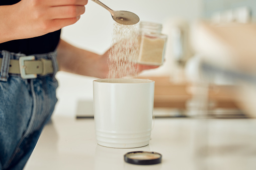 Sweet sugar pouring into mug of tea, coffee or hot drink at home. Closeup of woman making, drinking and preparing a warm beverage to enjoy with a teaspoon of substitute sweetener, glucose or stevia