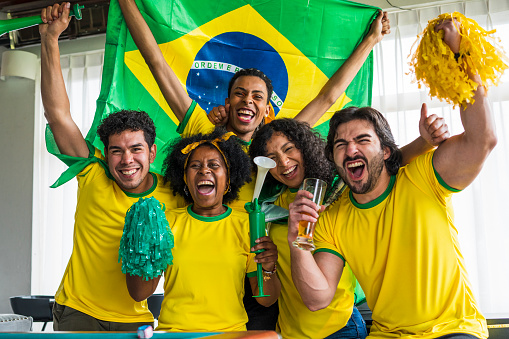 portraits of soccer fans, men and women from brazil, are at the house of one of them dressed in flags team jerseys, watching a soccer game while hugging each other and celebrating a big gollll