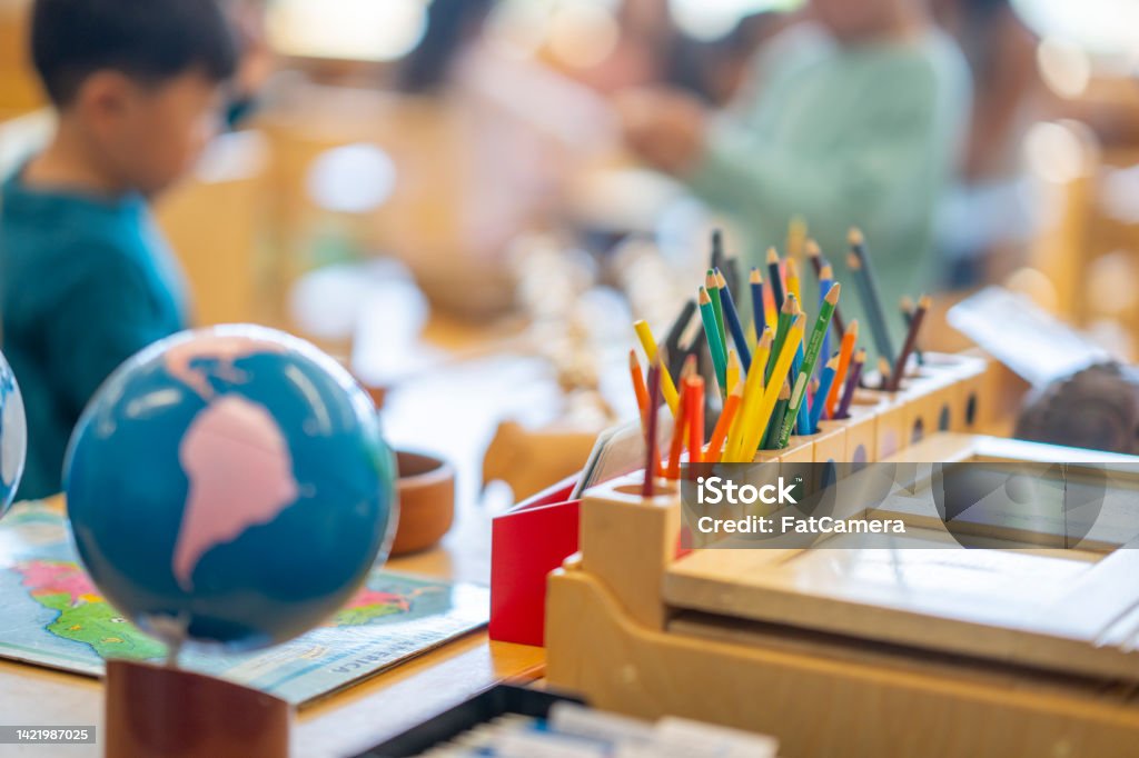 Montessori Classroom Set-Up A Montessori Geography classroom set-up complete with a world map, globes and pencil crayons all sitting out on a desk.  Two young students can be seen blurred in the background working away. Montessori Education Stock Photo