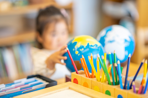 A Montessori Geography classroom set-up complete with a world map, globes and pencil crayons all sitting out on a desk.  A little girl can be seen reaching for one of the globes as she takes it off the shelf.