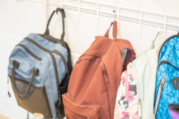 Children's backpacks are seen hanging up in a coatroom in a Montessori classroom.  They are various colors and patters and look very full.