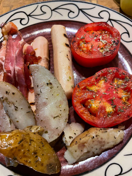 Stock photo showing a plate of chicken sausages, slices of bacon, fried tomato halves sprinkled with dry herbs and potato wedges, which is traditionally known as a fry-up. The components of a fry-up can vary from region to region. imagen de fotograma completo de plato con borde estampado con ingredientes fritos para el desayuno, salchichas de pollo cocidas, salchichas de tocino, mitades de tomate espolvoreadas con hierbas secas y rodajas de papa, enfoque en primer plano, vista eleva