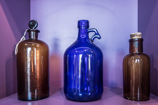 Glass bottles of shades of green, blue and amber rest on a shelf behind an outside dirty window of a Cape Cod antique shop.