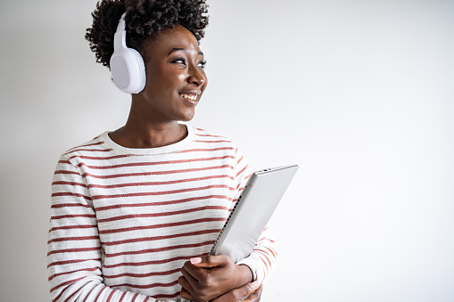 Young African American woman with wireless headphones holding laptop. She is standing against white wall and looking away.
