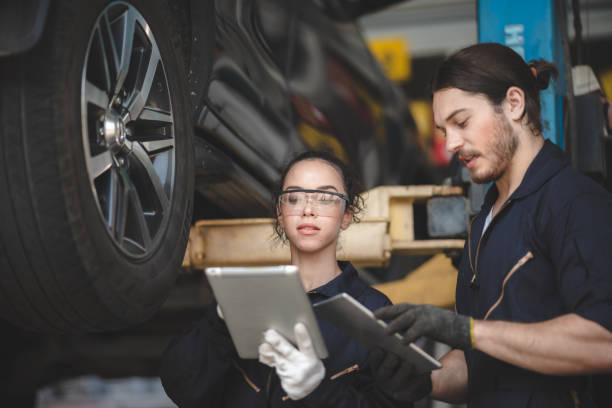 junge kfz-mechanikerin in uniform mit schutzbrille und handhandschuhen, die auf einem digitalen tablet arbeiten, während sie probleme im auto diagnostizieren - automechaniker stock-fotos und bilder