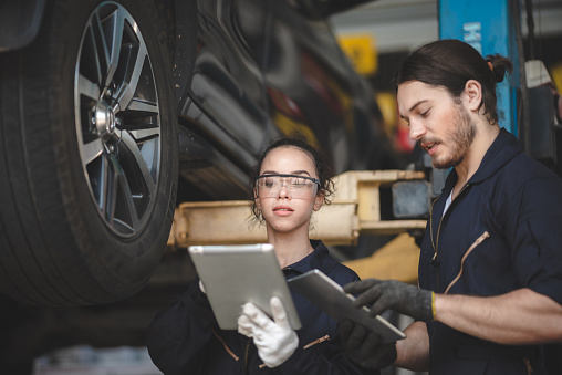 Young male and female car mechanic wearing uniform with safety eye glasses and hand gloves working on digital tablet while diagnosing problem in car in garage and discussing