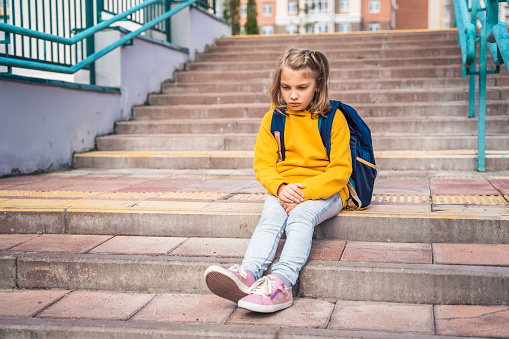 Back to elementary, primary school. Little sad unhappy girl with backpack. Lonely schoolgirl with emotional problems, victim of bullying in schoolyard. Teen in depresiion sitting alone on stair steps.