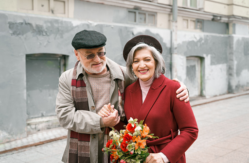 Elderly senior love couple. Old retired man woman together on romantic date.Aged husband wife walking on city street with flowers.Stylish elder hugging people pensioner in red coat.Happy family years.