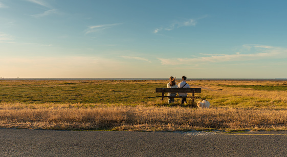 People (couple) sit on a bench in the middle of nowhere. Flat, peaceful, meadow landscape. Dunes, sea in the background. Brittany - North of France. English Channel (La Manche) region.