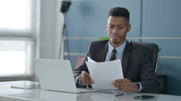 istock African Businessman with Laptop Reading Documents in Office 1422004934