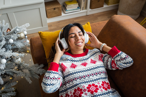 Young woman at home for Christmas. She is laying down on sofa and listening music on headphones.
