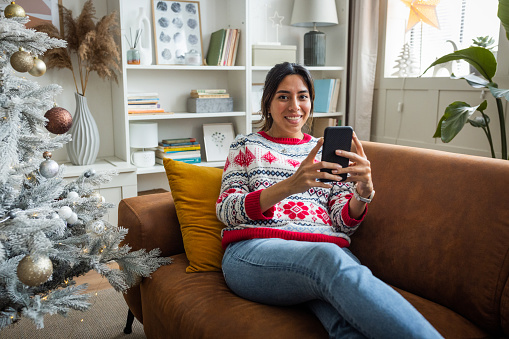 Young woman at home for Christmas. She is using smart phone while sitting on sofa.