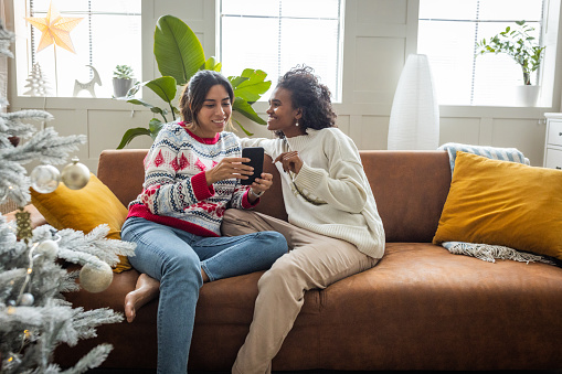 Female gay couple celebrating Christmas at home. They are sitting on sofa and using smart phone for shopping or pleasure.
