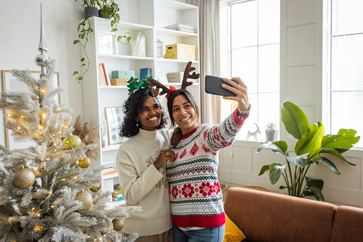 Female gay couple celebrating Christmas at home. They are decorating the Christmas tree and making selfie or photographing each other.