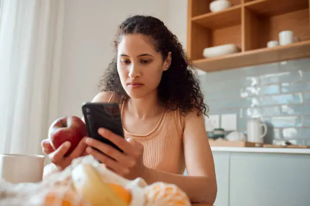 Photo of Eating healthy, diet planning and personal online female dietitian. Serious nutritionist check phone, snack on fresh fruit and concentrating on social media. Communicate with patient about calories.