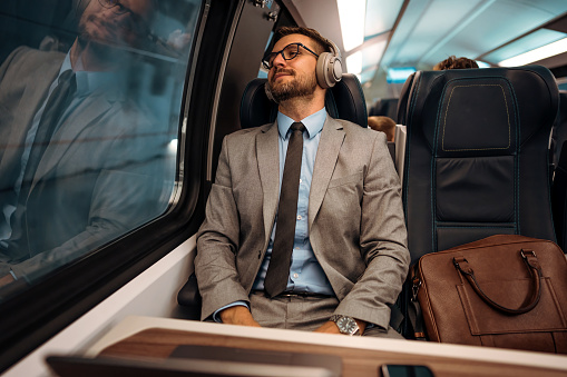 Tired handsome businessman relaxing while traveling with high speed train or metro. He is using laptop computer and wireless headphones for entertainment and music listening. Modern and fast travel concept.