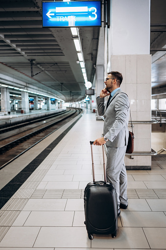 Handsome middle age businessman with suitcase standing in railroad station platform and waiting for high speed train or metro. Modern and fast travel concept.