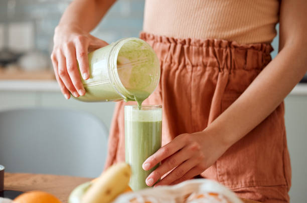 femme versant un smoothie sain dans un verre d’un pot mélangeur sur un comptoir pour la désintoxication. femme faisant du jus de fruits verts frais dans sa cuisine avec des légumes et des consommables pour un style de vie en forme. - juice drinking women drink photos et images de collection