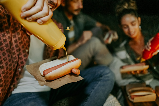 Friends enjoying hot dogs while camping outdoors at night, man pouring mustard over his hot dog