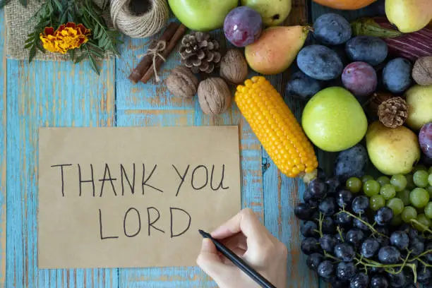Female hand writing note:"Thank You LORD" on vintage paper placed on wooden background with autumn fruit (grapes, plums, corn, apple, pumpkin). Christian gratitude, thanksgiving, and praise concept.