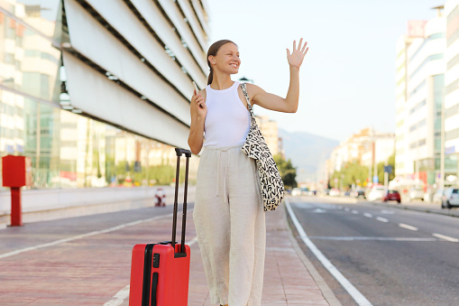 young beautiful woman with red suitcase with smartphone raising hand to catch taxi cab at terminal or station. stylish smiling girl tourist on summer holiday vacation. new trip.