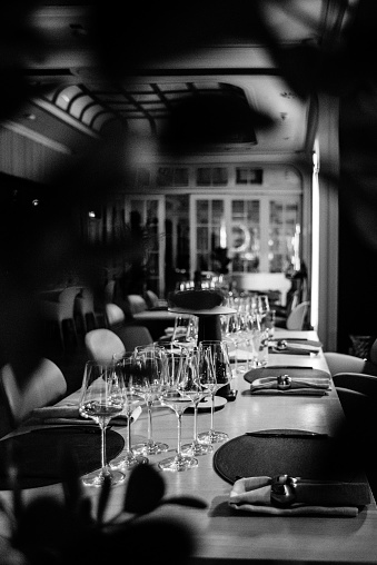 A black and white photograph of the interior of the restaurant through the leaves of a plant. Set long tables with appliances according to the rules of etiquette.
