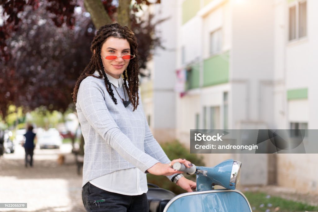 Hipster girl in pink sunglasses with dreadlocks standing near vintage motor scooter on city street Hipster girl in pink sunglasses with dreadlocks standing near vintage motor scooter on city street ready to drive 25-29 Years Stock Photo