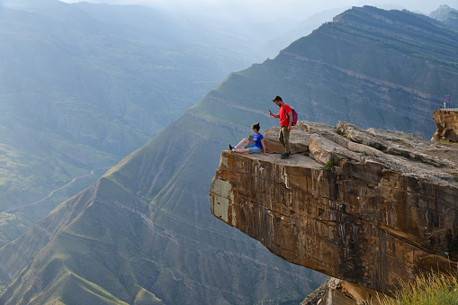 Dagestan, Russia - 21 July, 2022:  Young couple of tourists on the edge of cliff above deep abyss. Man takes a mobile photo oh his girlfriend. Ledges of rocks near the village of Goor