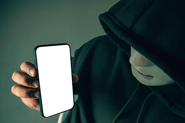 un hacker anonimo che indossa una maschera e una felpa nera con cappuccio mostra uno smartphone con uno schermo bianco vuoto. concetti di hacking e malware. - business stealing top secret confidential foto e immagini stock