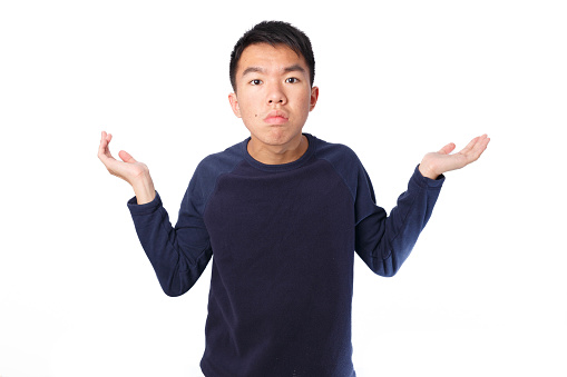 Medium shot of a young teen Asian boy in the studio with flash lighting. He is looking seriously at the camera, frowning and shrugging. He is wearing a navy blue sweater. The background is white.
