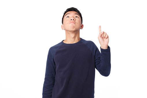 Medium shot of a young teen Asian boy in the studio with flash lighting. He is looking high up and pointing. He is wearing a navy blue sweater. The background is white.